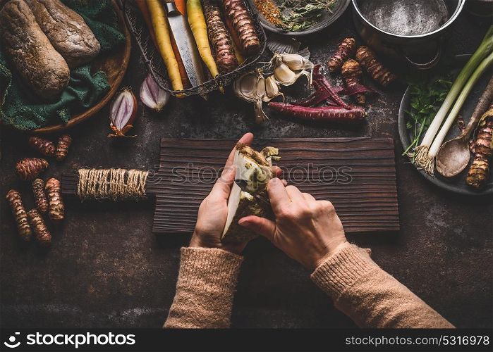 Female hands peeling celery on dark rustic kitchen table background with various vegetables and utensils. Root vegetables cooking preparation for tasty autumn dishes, top view. Healthy eating concept