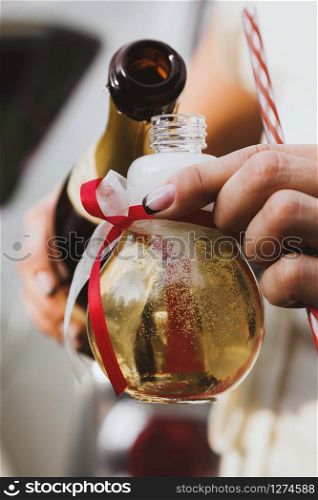 Female hands pouring champagne into a light bilb shaped party glass decorated with red and white ribbons
