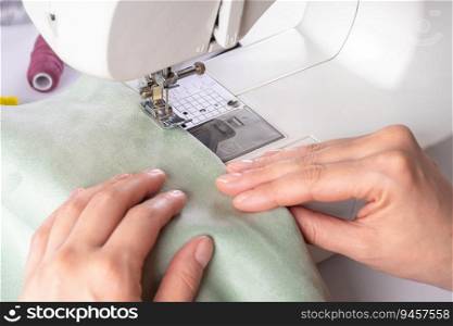 Female hands stitching green fabric on modern sewing machine at workplace in atelier. Women’s hands sew pieces of fabric on a sewing machine closeup. Handmade, hobby, small business concept