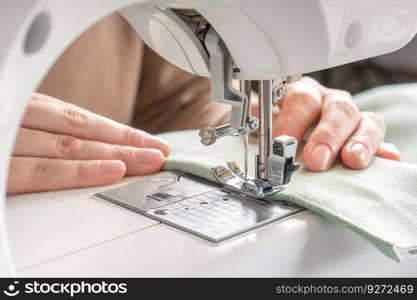 Female hands stitching white fabric on modern sewing machine at workplace in atelier. Women’s hands sew pieces of fabric on a sewing machine close-up. Handmade, hobby, small business concept