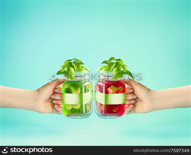 Female hands toasting with summer drinks. Red and green refreshing fruits beverages in glass jar with branding mock up at sunny bight mint turquoise background. Summer time mood. Blank Label . Health