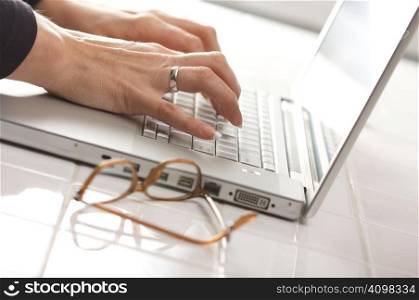 Female hands typing on the keyboard of the laptop with eyeglasses nearby.
