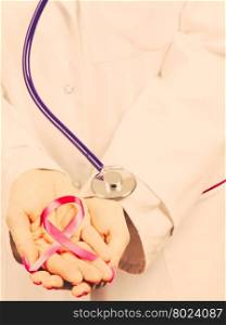Female hands with pink ribbon. Woman doctor with stethoscope holding pink cancer breast ribbon in hands. Awareness of being sick. Filtered.