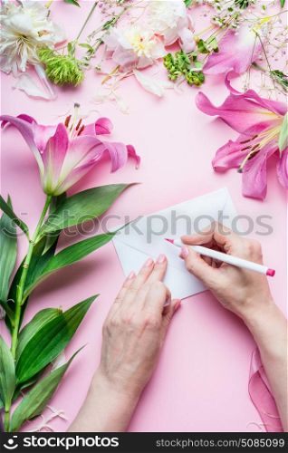 Female hands write with pencil greeting card on Blank envelop on pink table background with lily flowers and florist decoration equipment, top view. Creative Invitation and holiday concept