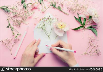 Female hands write with pencil on opened envelop with flowers arrangement . Florist decoration equipment on pink table background, top view. Invitation , greeting and holiday, concept