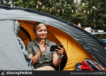 Female having video call with friends using smartphone while sitting in tent at camping. Woman relaxing in tent during summer vacation. Actively spending vacations outdoors close to nature. Concept of camp life