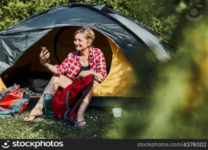 Female having video call with friends using smartphone while sitting in tent at camping. Woman relaxing in tent during summer vacation. Actively spending vacations outdoors close to nature. Concept of camp life