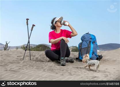 Female hiker sitting on sandy shore on Lanzarote and drinking hot beverage while having rest during trekking with cute Yorkshire Terrier dog. Female traveler drinking beverage during break on seashore