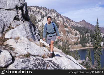 Female Hiker Walking Along A Steep Rocky Cliff Overlooking A Mountain Lake