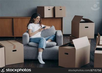 Female holding smartphone chooses relocation service or looking for repair ideas sitting in armchair with boxes. Smiling spanish woman tenant owner booking new dwelling on moving day.. Girl chooses relocation service, looks for repair ideas, using phone on moving day among boxes