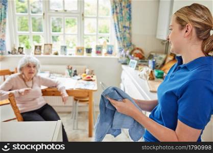 Female Home Help Cleaning House Doing Washing Up In Kitchen Whilst Chatting With Senior Woman