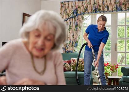 Female Home Help Cleaning House With Vacuum Cleaner Whilst Senior Woman Reads Newspaper