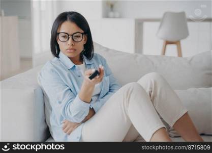 Female in glasses watching television, squinting, holding tv remote control, sitting on couch at home. Serious modern young woman changing channels, follows the news in live broadcast.. Female in glasses watching television squinting, holding tv remote control, sitting on couch at home