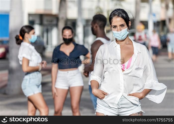 Female in summer outfit and protective mask standing on background of group of diverse friends while spending weekend on Lanzarote. Woman in mask standing against company of friends in city