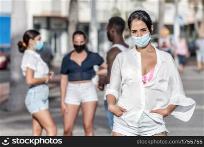 Female in summer outfit and protective mask standing on background of group of diverse friends while spending weekend on Lanzarote. Woman in mask standing against company of friends in city