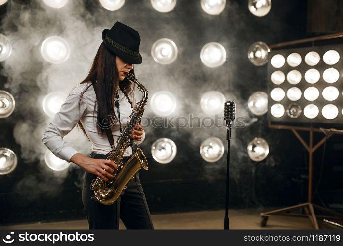 Female jazz musician in hat plays the saxophone on the stage with spotlights. Jazz performer playing on the scene. Female jazz musician in hat plays the saxophone