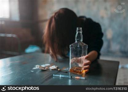 Female junkie with bottle of alcohol sitting at the table with drugs and syringe, grunge room interior on background. Drug addiction concept, addicted people. Junkie with bottle of alcohol sitting at the table