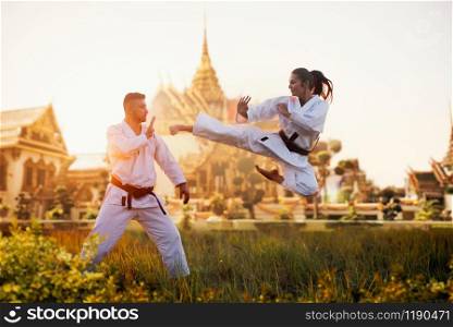 Female karate fighter trains kick in flight with master, training against ancient temple. Martial art fighters on workout outdoor, technique practice. Photo manipulation with background. Female karate fighter trains kick in flight