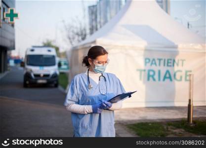 Female key doctor checking patient report form medical card,standing outside clinic exterior triage tent,Coronavirus COVID-19 illness diagnosis test,emergency due to global pandemic crisis,NHS UK EMS