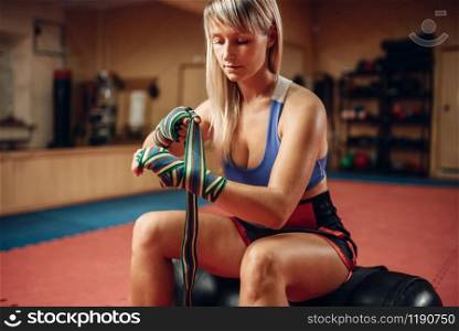 Female kickboxer in boxing bandages and sportswear sitting on punching bag after workout in the gym. Woman boxer on training, kickboxing practice. Female kickboxer sitting on punching bag in gym