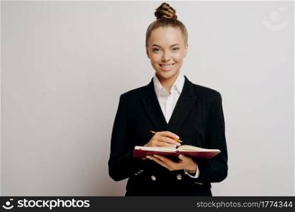 Female lawyer or business owner in dark suit with classic hairstyle writing down information about clients in red notebook with pencil while standing against light colored wall, smiling at camera. Business woman in dark suit writing in note book