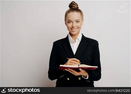 Female lawyer or business owner in dark suit with classic hairstyle, writing client info in red notebook, smiling at camera against light wall.