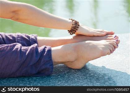 female legs in baggy cotton yoga trousers of purple color, hand holding her feet in stretching pose, outdoor, water in background. Healthy lifestyle, keep fit, weight loss concept