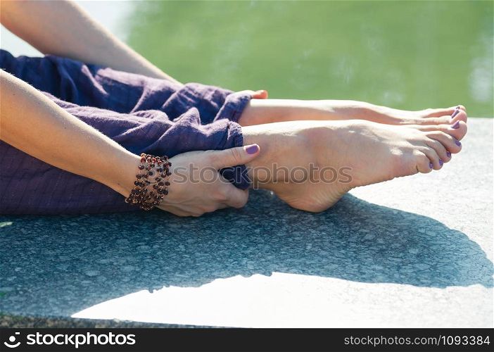 female legs in baggy cotton yoga trousers of purple color, hands holding her feet near ankles in stretching pose, outdoor, water in background. Healthy lifestyle, keep fit, weight loss concept