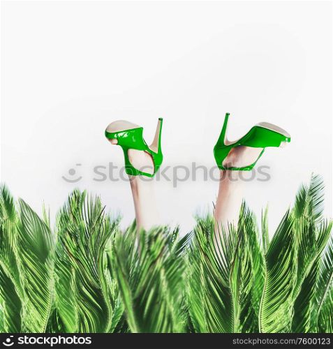 Female legs in green high heels shoes stick out from palm leaves at white background. Humorous summer time vacation concept. Fashion glamour stylish. Beauty