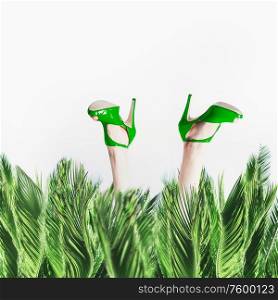 Female legs in green high heels shoes stick out from palm leaves at white background. Humorous summer time vacation concept. Fashion glamour stylish. Beauty