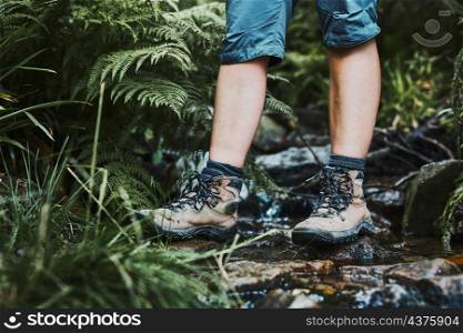 Female legs with trekking boots standing on stones in mountain stream. Summer vacation close to nature. Concept of actively spending leisure time