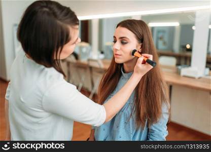 Female make up artist hand with brush applying make-up on beautiful girl face, beauty studio on background. Cosmetic salon. Female make up artist applying make-up with brush