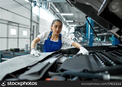 Female mechanic checks engine, car service. Vehicle repairing garage, woman in uniform, automobile station interior on background. Female mechanic checks engine, car service