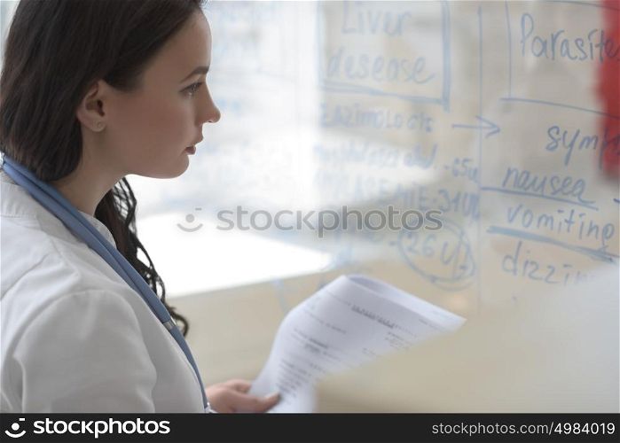 Female medical doctor working at clinic office. Writing on glass whiteboard symptoms and test results of her patient to diagnose disease