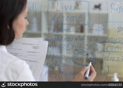 Female medical doctor working at clinic office. Writing on glass whiteboard symptoms and test results of her patient to diagnose disease