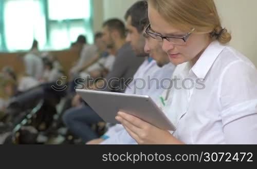 Female medical student is sitting in lecture hall during the break and chatting with friend using tablet PC.