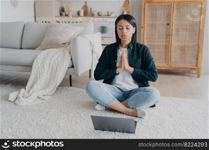 Female meditates to relieve stress, breathing deep, sitting in lotus pose at laptop on the floor at home. Calm woman in earphones listening audio affirmations, meditation does yoga. Healthy lifestyle.. Female meditates to relieve stress, breathing, sitting in lotus pose at laptop on the floor at home