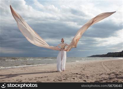 female model in extreme long beige dress posing dynamic in the beach. waving fabric. fluttering long skirt like a wings