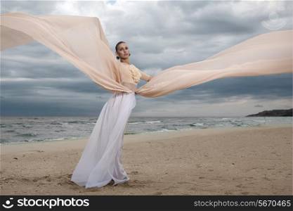 female model in long beige dress posing dynamic in the beach. waving fabric. fluttering long skirt