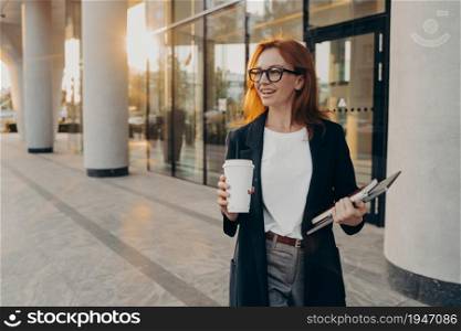 Female model with natural red hair dressed in formal wear holds touchpad notebook and cup of takeaway coffee thinks about content publication for sharing to website poses outdoor during sunny day. Female model holds touchpad notebook and cup of takeaway coffee poses outdoor during sunny day