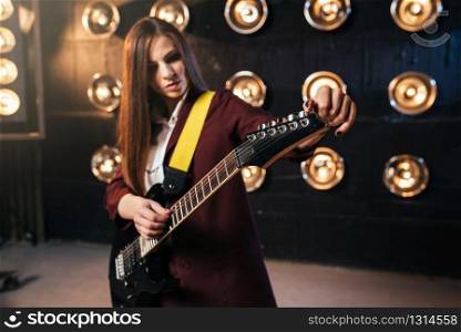 Female musician in suit playing on electric guitar, stage with lights on background, retro style. Live music performer, rock guitarist. Female musician in suit playing on electric guitar