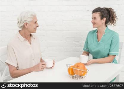 female nurse having coffee with senior woman sitting against white wall