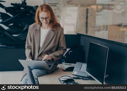 Female office worker in eyeglasses, wearing casual plaid jacket with jeans, sitting on top of desk in her cubicle, checking e-mail on laptop or working with project files before leaving office. Female office worker sitting on top of desk while working with project files in modern laptop