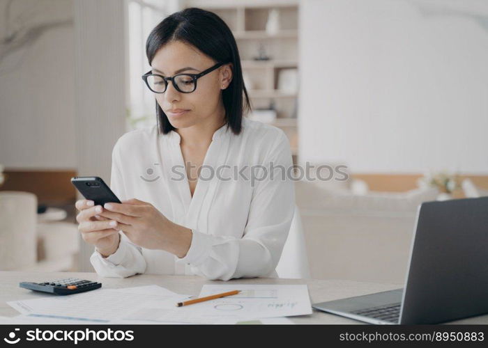 Female office worker sitting at laptop answer client’s message using mobile phone apps. Focused young businesswoman wearing glasses synchronize data between computer and smartphone.. Focused businesswoman in glasses answer client’s message using mobile phone apps sitting at laptop