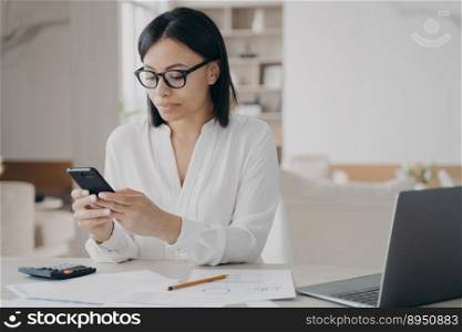 Female office worker sitting at laptop answer client’s message using mobile phone apps. Focused young businesswoman wearing glasses synchronize data between computer and smartphone.. Focused businesswoman in glasses answer client’s message using mobile phone apps sitting at laptop