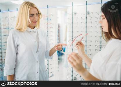 Female optician and buyer against showcase with glasses in optics store. Selection of eyeglasses with professional optometrist. Eye care, spectacles choice. Optician and buyer against showcase with glasses