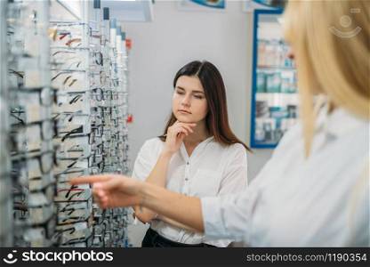 Female optician and buyer against showcase with glasses in optics store. Selection of eyeglasses with professional optometrist. Eye care, spectacles choice