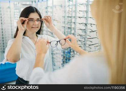 Female optician and buyer chooses glasses frame against showcase with eyeglasses in optics store. Selection of spectacles with professional optometrist