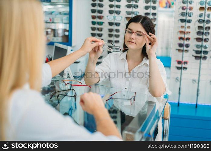 Female optician and consumer chooses glasses frame in optics store. Selection of spectacles with professional optometrist
