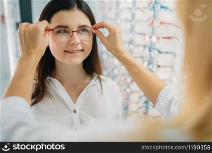 Female optician and customer chooses glasses frame against showcase with eyeglasses in optics store. Selection of spectacles with professional optometrist